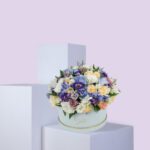 A round, light blue hat box filled with a large arrangement of pastel-colored flowers. The flowers include shades of purple, blue, white, and pink. The box is sitting on a white platform against a light pink background.