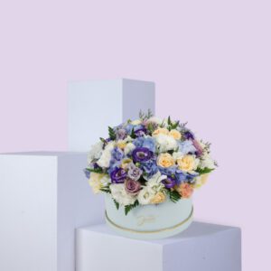 A round, light blue hat box filled with a large arrangement of pastel-colored flowers. The flowers include shades of purple, blue, white, and pink. The box is sitting on a white platform against a light pink background.
