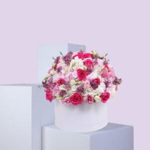 A round, white hat box filled with a large arrangement of pink and white flowers. The flowers include pink roses, white hydrangeas, and other pink and white blooms. The box is sitting on a white platform against a light pink background.