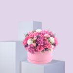 A round, pink hat box filled with a large arrangement of pink and white flowers. The flowers include pink roses, white hydrangeas, and other pink and white blooms. The box is sitting on a white platform against a light pink background.