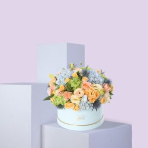 A round, light blue hat box filled with a large arrangement of pastel-colored flowers. The flowers include shades of purple, blue, white, and pink. The box is sitting on a white platform against a light pink background.