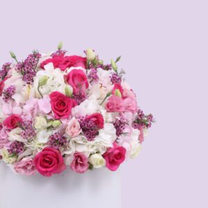 A round, white hat box filled with a large arrangement of pink and white flowers. The flowers include pink roses, white hydrangeas, and other pink and white blooms. The box is sitting on a white platform against a light pink background.