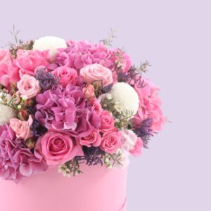 A round, pink hat box filled with a large arrangement of pink and white flowers. The flowers include pink roses, white hydrangeas, and other pink and white blooms. The box is sitting on a white platform against a light pink background.
