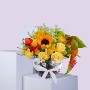 A vibrant bouquet of sunflowers, roses, and other colorful flowers arranged in a glass vase with a white ribbon.