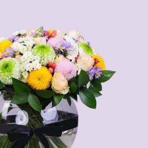 A colorful bouquet of flowers in a round glass vase, tied with a black ribbon. The bouquet includes chrysanthemums, roses, and other vibrant flowers.