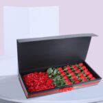 A black gift box open to reveal a compartment of red roses and another compartment of red rose petals.