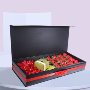 A black gift box open to reveal a compartment of red roses and another compartment of red rose petals. There is also a small gold gift bag in the box.