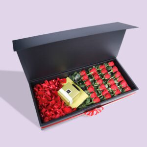 A black gift box open to reveal a compartment of red roses and another compartment of red rose petals. There is also a small gold gift bag in the box.