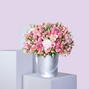 A round, silver hat box filled with a large arrangement of pink flowers, including roses, hydrangeas, and other pink blooms.