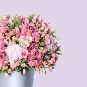 A round, silver hat box filled with a large arrangement of pink flowers, including roses, hydrangeas, and other pink blooms.