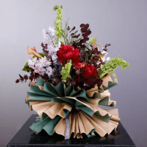 A vibrant flower arrangement in a unique paper wrapping. The bouquet features a mix of red, white, and green flowers, including roses, delphiniums, and other blooms. The paper wrapping is a combination of brown and green, creating a visually appealing contrast.