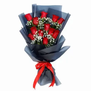 The image shows a bouquet of red roses arranged in a classic way, with the stems wrapped in black paper and a small red ribbon tied around the base. The bouquet is sitting on a white pedestal against a light gray background.