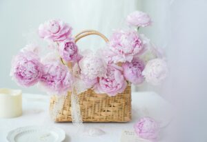 The image shows a beautiful arrangement of pink peonies in a woven basket. The basket is placed on a white table with a plate and a candle. The overall aesthetic is soft, feminine, and romantic.