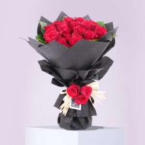The image shows a beautiful bouquet of red roses. The roses are arranged in a classic way, with the stems wrapped in black paper and a small bow of red roses tied around the base. The bouquet is sitting on a white pedestal against a light gray background.