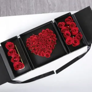 The image shows a rectangular black box with three compartments. The box is open, revealing red roses arranged in a heart shape in the center compartment and two smaller arrangements of red roses on either side. The arrangement spells out the words "I Love You."