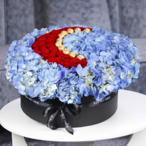 The image shows a large, round flower arrangement in a black box. The arrangement is made up of red roses, blue hydrangeas, and Ferrero Rocher chocolates.