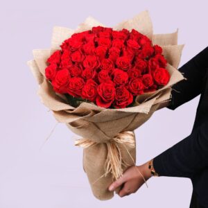 The image shows a large bouquet of red roses wrapped in burlap. The burlap is tied with a raffia ribbon.