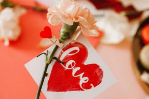 Close-up of a peach carnation with a heart-shaped card that says "love" in cursive, clipped to the stem.