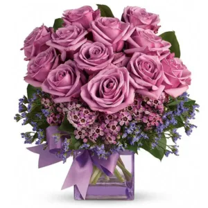 A bouquet of purple roses in a square glass vase with purple ribbon and blue accent flowers.