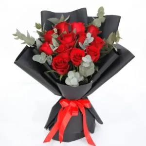 Red roses and eucalyptus leaves arranged in a bouquet with black wrapping and a red bow.
