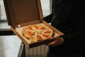 he person's hands are holding the box, presenting the fresh pizza. The blurred background shows an indoor environment, possibly a home or office setting, with a glimpse of a light-colored surface and a window with vertical blinds, further implying the pizza has just been brought inside